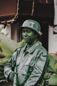 A person dressed as a toy soldier with green paint covering their face and uniform. The attire includes a helmet and a military-style jacket. The background features large green leaves, adding to the camouflage effect.