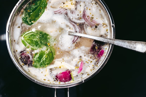 A pot filled with a bubbling liquid containing bright green spinach leaves, slices of red onions, and opaque chunks of mushroom or tofu. The silver spoon resting inside suggests ongoing cooking or preparation. Tiny spices are scattered across the surface, indicating a savory dish.