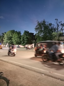 The image captures a street scene at dusk with blurred motion of motorcycles and a bicycle on the sidewalk. Trees and a building are visible in the background, with the sky transitioning to a darker shade as evening approaches.