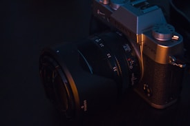 A vintage-style digital camera with a textured grip and a large lens barrel. The camera body features dials and brand markings, and it's surrounded by a soft, warm lighting against a dark background.