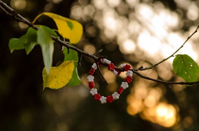 A handmade heart-shaped bracelet made of red and white beads is delicately hanging on a thin branch. Surrounding it are green leaves with hints of yellow, and the background is filled with a soft, glowing bokeh effect, creating a warm and nostalgic ambiance.
