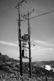 A wooden electrical pole stands against a clear sky with clouds, featuring numerous wires, insulators, and a transformer. The foreground includes a rocky, grassy hillside, while a white building and distant hills are visible in the background.