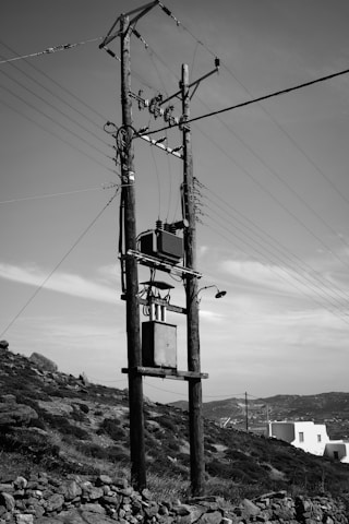 A wooden electrical pole stands against a clear sky with clouds, featuring numerous wires, insulators, and a transformer. The foreground includes a rocky, grassy hillside, while a white building and distant hills are visible in the background.
