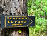 A wooden sign attached to a tree shows a directional arrow with the text 'Sendero El Puma (Dist 600 mts)' in bright yellow lettering with a forest in the background. A small yellow bottle cap is embedded into the tree below the sign.