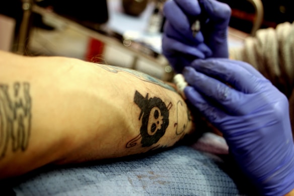 A person is getting a tattoo on their arm, with a design featuring a black skull. The tattoo artist is wearing blue gloves and is actively working on the piece using tattoo equipment. The environment suggests a professional setting with a focus on the arm and tattoo.