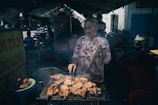 An older woman stands at a grill, cooking meat skewers and chicken outdoors in a bustling market area. She wears a patterned blouse and holds cooking utensils, surrounded by plastic bowls and signs with text. The background features other market stalls and motorbikes, indicating a lively atmosphere.
