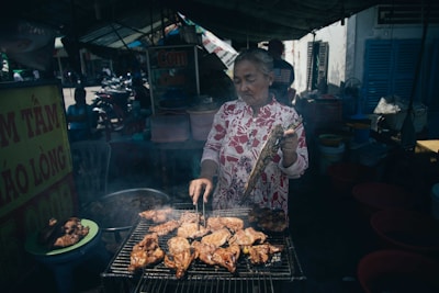 An older woman stands at a grill, cooking meat skewers and chicken outdoors in a bustling market area. She wears a patterned blouse and holds cooking utensils, surrounded by plastic bowls and signs with text. The background features other market stalls and motorbikes, indicating a lively atmosphere.
