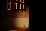 A dimly lit interior with a neon 'BAR' sign glowing in red against a brick wall. Below the sign, there is a tiled wall and a countertop. On the counter, a glass with a napkin can be seen, along with a yellow object that resembles a condiment container.
