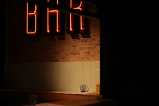 A dimly lit interior with a neon 'BAR' sign glowing in red against a brick wall. Below the sign, there is a tiled wall and a countertop. On the counter, a glass with a napkin can be seen, along with a yellow object that resembles a condiment container.