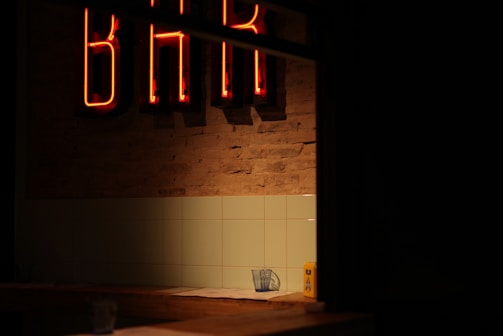 A dimly lit interior with a neon 'BAR' sign glowing in red against a brick wall. Below the sign, there is a tiled wall and a countertop. On the counter, a glass with a napkin can be seen, along with a yellow object that resembles a condiment container.