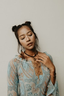 A young person with their eyes closed, wearing an intricately patterned blue and brown outfit. The individual has neatly styled hair in two buns and is adorned with white facial markings. They have a calm expression and their hand gently touches their collar.