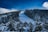 A snow-covered mountain slope with a ski run stretches through a dense forest of evergreen trees. Several wooden cabins with steep roofs are nestled among the tall trees. The sky above is a vibrant blue with some clouds, adding contrast to the snowy landscape.