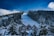 A snow-covered mountain slope with a ski run stretches through a dense forest of evergreen trees. Several wooden cabins with steep roofs are nestled among the tall trees. The sky above is a vibrant blue with some clouds, adding contrast to the snowy landscape.