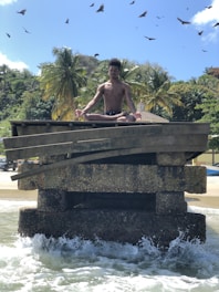 A person is meditating in a lotus position on a weathered concrete structure surrounded by ocean waves. The background features lush palm trees and a few buildings, set against a clear blue sky with birds soaring overhead.