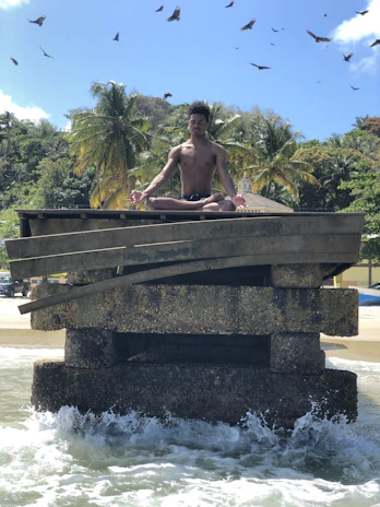A person is meditating in a lotus position on a weathered concrete structure surrounded by ocean waves. The background features lush palm trees and a few buildings, set against a clear blue sky with birds soaring overhead.