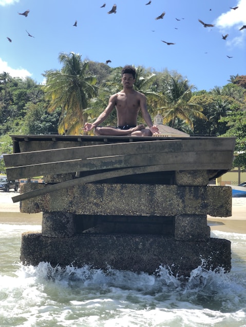 A person is meditating in a lotus position on a weathered concrete structure surrounded by ocean waves. The background features lush palm trees and a few buildings, set against a clear blue sky with birds soaring overhead.
