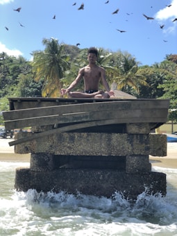 A person is meditating in a lotus position on a weathered concrete structure surrounded by ocean waves. The background features lush palm trees and a few buildings, set against a clear blue sky with birds soaring overhead.