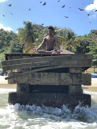 A person is meditating in a lotus position on a weathered concrete structure surrounded by ocean waves. The background features lush palm trees and a few buildings, set against a clear blue sky with birds soaring overhead.