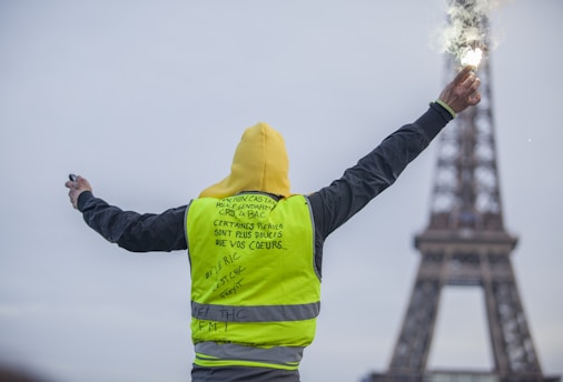 A person wearing a yellow vest stands with arms outstretched, holding a flare in one hand. The Eiffel Tower is visible in the background, slightly blurred, indicating a location in Paris. The text on the vest includes messages written in French.