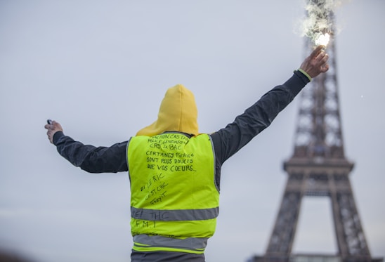 A person wearing a yellow vest stands with arms outstretched, holding a flare in one hand. The Eiffel Tower is visible in the background, slightly blurred, indicating a location in Paris. The text on the vest includes messages written in French.