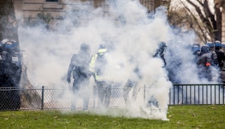 Several individuals wearing protective gear are surrounded by a cloud of smoke or tear gas in an outdoor setting. They appear to be part of a larger group near a fence, with some trees and buildings visible in the background.