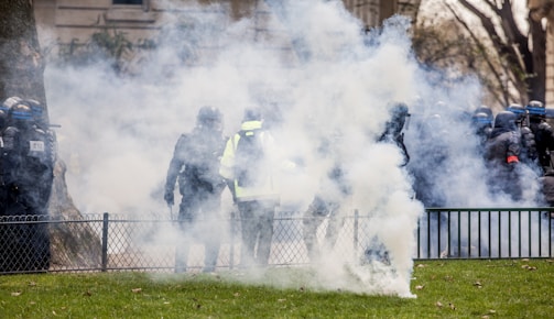 Several individuals wearing protective gear are surrounded by a cloud of smoke or tear gas in an outdoor setting. They appear to be part of a larger group near a fence, with some trees and buildings visible in the background.
