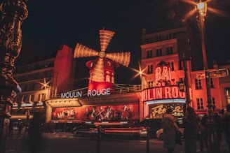 A vibrant view of a bustling street at night, featuring a well-lit entertainment venue with a large, iconic windmill and neon lights. The building is adorned with red and orange tones, and there's a crowd of people nearby along with moving vehicles.