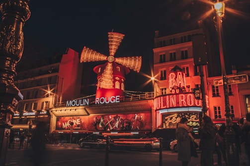 A vibrant view of a bustling street at night, featuring a well-lit entertainment venue with a large, iconic windmill and neon lights. The building is adorned with red and orange tones, and there's a crowd of people nearby along with moving vehicles.