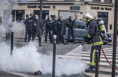 Firefighter in protective gear is using a hose to spray water on smoke emerging from the ground in an urban setting. A group of police officers in full riot gear stand in the background, creating a tense and controlled atmosphere.