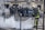 Firefighter in protective gear is using a hose to spray water on smoke emerging from the ground in an urban setting. A group of police officers in full riot gear stand in the background, creating a tense and controlled atmosphere.