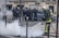 Firefighter in protective gear is using a hose to spray water on smoke emerging from the ground in an urban setting. A group of police officers in full riot gear stand in the background, creating a tense and controlled atmosphere.