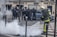 Firefighter in protective gear is using a hose to spray water on smoke emerging from the ground in an urban setting. A group of police officers in full riot gear stand in the background, creating a tense and controlled atmosphere.