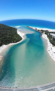 A scenic coastal area with a river flowing into the ocean, bordered by sandy beaches and dense green vegetation. The water displays a gradient from deep blue in the ocean to turquoise and lighter shades in the river. Several people are visible on the beach and in the water, enjoying recreational activities.