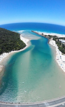 A scenic coastal area with a river flowing into the ocean, bordered by sandy beaches and dense green vegetation. The water displays a gradient from deep blue in the ocean to turquoise and lighter shades in the river. Several people are visible on the beach and in the water, enjoying recreational activities.