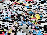 A pile of interlocking jigsaw puzzle pieces with a variety of colors and illustrations, some depicting characters or scenes from a comic or cartoon style. The pieces are scattered randomly, showing partial sections of an image.