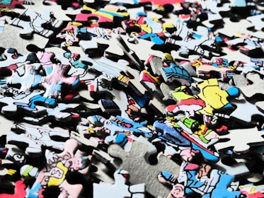 A pile of interlocking jigsaw puzzle pieces with a variety of colors and illustrations, some depicting characters or scenes from a comic or cartoon style. The pieces are scattered randomly, showing partial sections of an image.