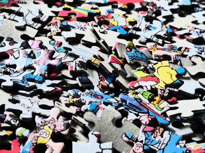 A pile of interlocking jigsaw puzzle pieces with a variety of colors and illustrations, some depicting characters or scenes from a comic or cartoon style. The pieces are scattered randomly, showing partial sections of an image.