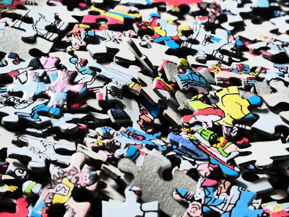 A pile of interlocking jigsaw puzzle pieces with a variety of colors and illustrations, some depicting characters or scenes from a comic or cartoon style. The pieces are scattered randomly, showing partial sections of an image.