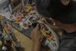 A person is customizing a shoe with intricate designs and colorful patterns. Various paint pots and brushes are scattered on a wooden table, alongside a smartphone displaying a design. The person is wearing a cap.