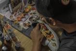 A person is customizing a shoe with intricate designs and colorful patterns. Various paint pots and brushes are scattered on a wooden table, alongside a smartphone displaying a design. The person is wearing a cap.