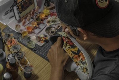 A person is customizing a shoe with intricate designs and colorful patterns. Various paint pots and brushes are scattered on a wooden table, alongside a smartphone displaying a design. The person is wearing a cap.