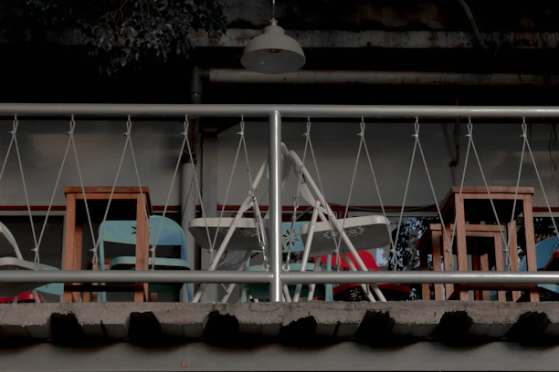 A balcony scene includes a series of foldable chairs and wooden stools arranged behind a metal railing with a criss-cross design. There are colorful abstract patterns on a mural in the background, and a lamp hangs above the space, casting shadows.