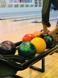 A selection of colorful bowling balls is seen in a bowling alley with someone's hands selecting a ball. The background shows multiple bowling lanes and a clear, smooth wooden floor.
