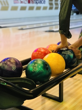 A selection of colorful bowling balls is seen in a bowling alley with someone's hands selecting a ball. The background shows multiple bowling lanes and a clear, smooth wooden floor.