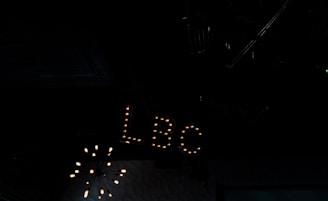 A dimly lit interior space featuring a vintage-style sign with the letters 'LBC' illuminated by small light bulbs. The background is dark, with a chandelier-like light fixture partially visible above.