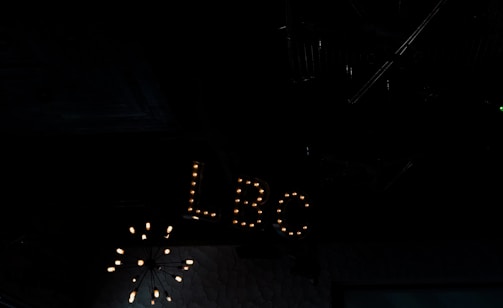 A dimly lit interior space featuring a vintage-style sign with the letters 'LBC' illuminated by small light bulbs. The background is dark, with a chandelier-like light fixture partially visible above.