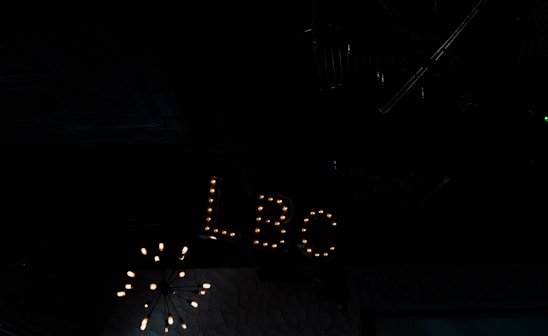 A dimly lit interior space featuring a vintage-style sign with the letters 'LBC' illuminated by small light bulbs. The background is dark, with a chandelier-like light fixture partially visible above.
