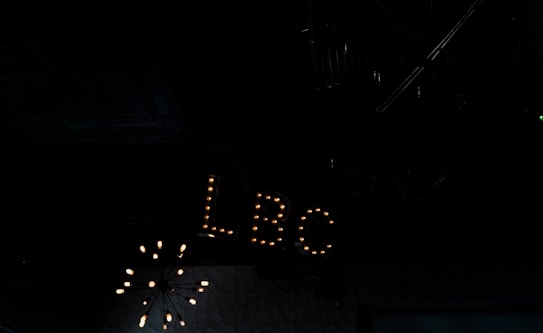 A dimly lit interior space featuring a vintage-style sign with the letters 'LBC' illuminated by small light bulbs. The background is dark, with a chandelier-like light fixture partially visible above.