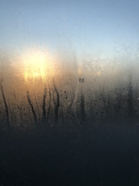 Condensation on a window with streaks running down. Behind the window, there's a bright light source, likely the sun, creating a diffused glow through the moisture. The sky in the background is clear and changes from blue at the top to a lighter hue near the horizon.
