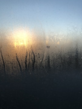 Condensation on a window with streaks running down. Behind the window, there's a bright light source, likely the sun, creating a diffused glow through the moisture. The sky in the background is clear and changes from blue at the top to a lighter hue near the horizon.